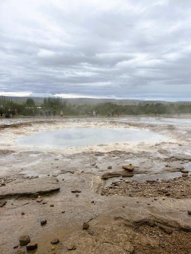 La palabra géiser procede de ‘Geysir’, que proviene del verbo islandés del nórdico antiguo ‘geysa’, que significa ‘salir a chorro’. La actividad de los géiseres está provocada por el contacto entre el agua superficial y las rocas calentadas por el magma. El agua que se calienta bajo la tierra en cámaras magmáticas regresa a la superficie a través de conductos o estrechas grietas formadas en rocas fracturadas. A medida que estos depósitos se llenan, el agua más profunda se calienta y la más superficial se va enfriando, pero debido a lo estrecho del conducto, el enfriamiento del agua en profundidad es imposible. El agua caliente presiona desde abajo al agua fría de la superficie, semejante a la tapa de una olla a presión, haciendo que el agua del depósito se sobrecaliente y supere el punto de ebullición (sobre 120º C) aumentando la presión, lo que provoca que ascienda el vapor de agua por el conducto y salga violentamente a la superficie formando una columna de agua y vapor de agua.
En la imagen el Geiser Strokkur. (Autor: Antonio P. López)