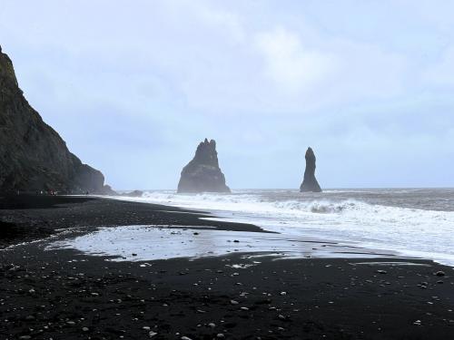 Al este de los acantilados de Reynisfjall, se encuentra la playa de ‘arena negra’ de Reynisfjara, en Vík í Mýrdal. La playa está formada por pequeños fragmentos negros y brillantes de basalto, de unos 2-3 mm, como producto del rápido enfriamiento del magma en contacto con el agua, que hace que se descomponga en trozos más pequeños, y de la fuerte erosión marina posterior. Además de basalto, la arena negra está formada por restos de piroxenos (augita), olivino, plagioclasa (labradorita), magnetita e ilmenita, en cantidades variables.
En la imagen, dos agujas de basalto en la playa de Reynisfjara, conocidas como Reynisdrangar. Miden 43 metros de altura y son el resultado de la erosión del material más blando que rodeaba al basalto más duro. Para los que vieron la serie de HBO ’Juego de Tronos’, Reynisdrangar aparece en un capítulo de la séptima temporada. (Autor: Antonio P. López)