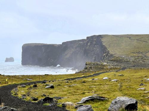 Los acantilados de Reynisfjall, cerca del pueblo de Vík í Mýrdal, al sur de Islandia, se formaron a partir de una fisura submarina en el Cinturón Volcánico Oriental, en el sistema volcánico Katla, a finales del Pleistoceno. Con una altitud de 120 metros, están constituidos por una alternancia de rocas ígneas extrusivas e intrusivas que indican que se formaron en varias fases eruptivas. En la base aparecen ‘pillow lavas’ y columnas de composición basáltica, seguidas de tobas volcánicas, a menudo con intrusiones, y en la parte superior capas de lava. Originalmente era una isla antes de convertirse en una península, unida al continente en el Holoceno, por la combinación de procesos fluviales, glaciares, volcánicos y marinos.
En la base de los acantilados aparecen unos arcos que se formaron a partir de cuevas marinas y por la acción erosiva de las olas. El más conocido es el Dyrhólaey. (Autor: Antonio P. López)