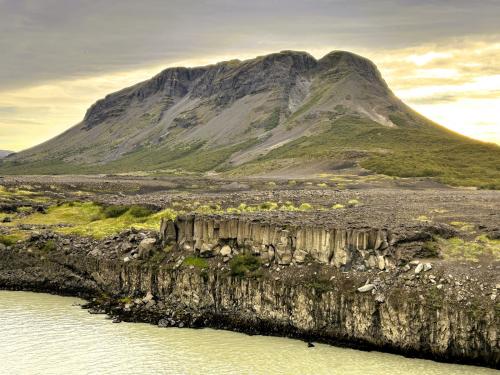 El monte Búrfell, un tipo de volcán denominado ‘tuya’, de 480 metros de altura está al norte de Þjófafoss. Un ‘tuya’ es un tipo de volcán que se forma cuando entra en erupción bajo un glaciar o bajo potentes capas de hielo. Este tipo de volcanes se reconocen por estar formados por capas casi horizontales de rocas basálticas y laderas escarpadas. El magma en erupción bajo un glaciar se enfría con relativa rapidez y no se desplaza muy lejos, por lo que se concentra formando una colina de laderas escarpadas con una cima plana. Este tipo de montañas son bastante raras en todo el mundo, ya que se limitan a zonas en las que el vulcanismo activo se produce al mismo tiempo que la cobertura glaciar. El origen del término proviene de ‘Tuya Butte’, una de las muchas tuyas en la zona del río Tuya y la cordillera Tuya (Tuya Range) en el extremo norte de la Columbia Británica, en Canadá.
En la parte inferior de la fotografía se aprecian alineaciones de columnas basálticas en el cauce del río Þjórsá. (Autor: Antonio P. López)