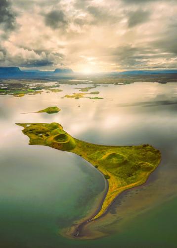 El lago Mývatn, en el municipio de Skútustaðahreppur, al norte de Islandia, con 37 km2 de superficie y 4,5 metros de profundidad máxima, alberga una serie de curiosos cráteres que afloran en su superficie, son los Skútustaðagígar. La traducción al castellano es literalmente "cráteres de Skútustaðir", y son un conjunto de ‘pseudocráteres’ situados en la costa sur del lago. El apelativo de ‘pseudocráteres’ se lo dio el geólogo y vulcanólogo islandés Sigurður Þórarinsson porque nunca llegaron a emitir lava. Según su teoría, Skútustaðir se creó hace aproximadamente 2.300 años, durante la erupción simultánea del Lúdentaborgir y el Þrengslaborgir, del sistema volcánico Krafla. En su avance, la lava alcanzó una zona pantanosa o un lago somero muy parecido al actual Mývatn. El enorme calor evaporó casi inmediatamente el agua superficial, pero aquella que se encontraba a mayor profundidad se fue calentando más lentamente. Mientras tanto, el peso de la lava acumulada en la superficie aumentaba la presión sobre la mezcla de sedimentos y agua. Finalmente, el agua acabó abriéndose camino en forma de vapor a través de la colada en una serie de erupciones explosivas que formaron estos conos.
Foto tomada con dron por Carlos Martín Ayala (Autor: Antonio P. López)