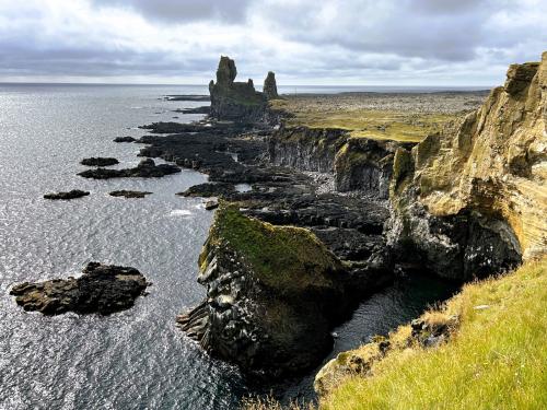 En la parte más meridional de la costa de la península de Snæfellsnes, en Breiðavíkurhreppur y dentro del Parque Nacional Snæfellsjökull, se encuentra el acantilado de Þúfubjarg (a la derecha de la imagen). Está constituido por capas de toba volcánica de color ocre, formadas por la acumulación de cenizas y lapilli durante erupciones piroclásticas violentas. Estos depósitos de toba submarinos fueron erosionados por el mar dejando a la vista las capas internas del cono volcánico.
Al fondo, aparecen dos pináculos de basalto, Lóndrangar, que son los restos de un antiguo domo o tapón de un edificio volcánico mayor que ha sido erosionado. La aguja mayor mide 75 metros de altura, mientras que la menor mide 61 metros. El origen podría estar en que el magma basáltico se solidificó dentro de una chimenea secundaria o respiradero y luego quedó expuesto a la vista por la erosión del material circundante más blando. (Autor: Antonio P. López)