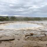 La palabra géiser procede de ‘Geysir’, que proviene del verbo islandés del nórdico antiguo ‘geysa’, que significa ‘salir a chorro’. La actividad de los géiseres está provocada por el contacto entre el agua superficial y las rocas calentadas por el magma. El agua que se calienta bajo la tierra en cámaras magmáticas regresa a la superficie a través de conductos o estrechas grietas formadas en rocas fracturadas. A medida que estos depósitos se llenan, el agua más profunda se calienta y la más superficial se va enfriando, pero debido a lo estrecho del conducto, el enfriamiento del agua en profundidad es imposible. El agua caliente presiona desde abajo al agua fría de la superficie, semejante a la tapa de una olla a presión, haciendo que el agua del depósito se sobrecaliente y supere el punto de ebullición (sobre 120º C) aumentando la presión, lo que provoca que ascienda el vapor de agua por el conducto y salga violentamente a la superficie formando una columna de agua y vapor de agua.
En la imagen el Geiser Strokkur. (Autor: Antonio P. López)