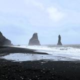 Al este de los acantilados de Reynisfjall, se encuentra la playa de ‘arena negra’ de Reynisfjara, en Vík í Mýrdal. La playa está formada por pequeños fragmentos negros y brillantes de basalto, de unos 2-3 mm, como producto del rápido enfriamiento del magma en contacto con el agua, que hace que se descomponga en trozos más pequeños, y de la fuerte erosión marina posterior. Además de basalto, la arena negra está formada por restos de piroxenos (augita), olivino, plagioclasa (labradorita), magnetita e ilmenita, en cantidades variables.
En la imagen, dos agujas de basalto en la playa de Reynisfjara, conocidas como Reynisdrangar. Miden 43 metros de altura y son el resultado de la erosión del material más blando que rodeaba al basalto más duro. Para los que vieron la serie de HBO ’Juego de Tronos’, Reynisdrangar aparece en un capítulo de la séptima temporada. (Autor: Antonio P. López)