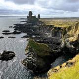 En la parte más meridional de la costa de la península de Snæfellsnes, en Breiðavíkurhreppur y dentro del Parque Nacional Snæfellsjökull, se encuentra el acantilado de Þúfubjarg (a la derecha de la imagen). Está constituido por capas de toba volcánica de color ocre, formadas por la acumulación de cenizas y lapilli durante erupciones piroclásticas violentas. Estos depósitos de toba submarinos fueron erosionados por el mar dejando a la vista las capas internas del cono volcánico.
Al fondo, aparecen dos pináculos de basalto, Lóndrangar, que son los restos de un antiguo domo o tapón de un edificio volcánico mayor que ha sido erosionado. La aguja mayor mide 75 metros de altura, mientras que la menor mide 61 metros. El origen podría estar en que el magma basáltico se solidificó dentro de una chimenea secundaria o respiradero y luego quedó expuesto a la vista por la erosión del material circundante más blando. (Autor: Antonio P. López)